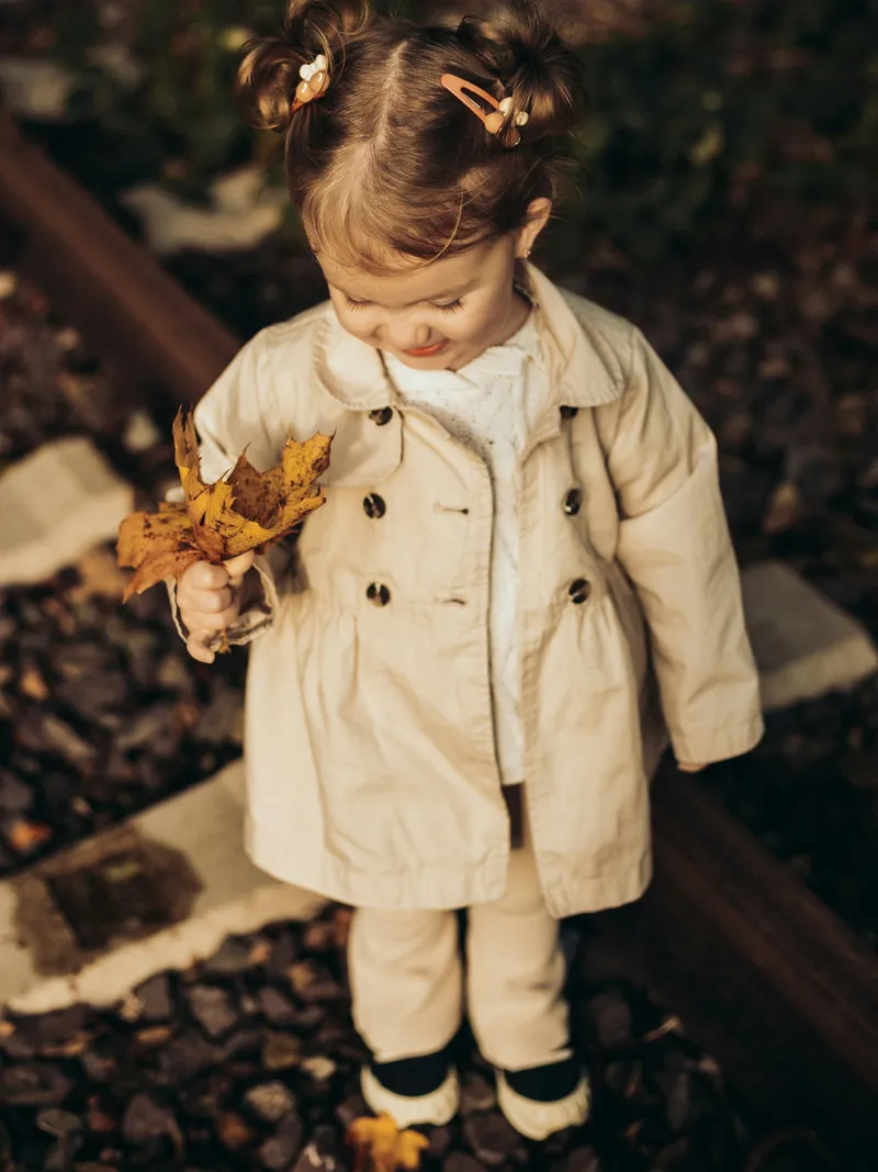 Toddler girl in a stone double-breasted trench coat holding autumn leaves