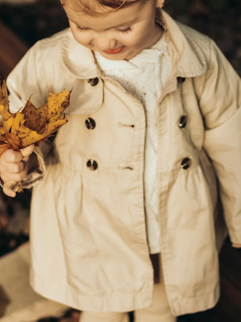 Toddler girl in a stone double-breasted trench coat holding autumn leaves