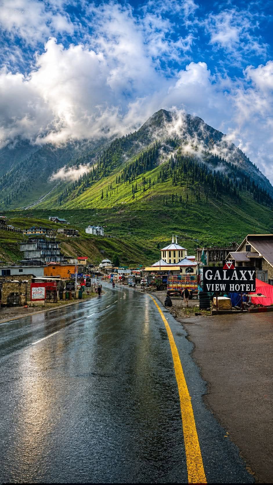 Rain-washed road leading into Naran bazaar with a cloud-wrapped green peak rising behind the town